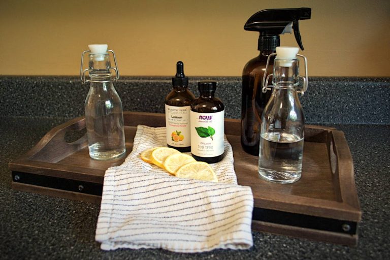 Wooden serving board with a cloth napkin on top, freeze-dried lemons, lemon essential oil, tea tree essential oil, two clear swing-top bottles with liquid and an amber-colored spray bottle. These are ingredients to make all-purpose cleaner.