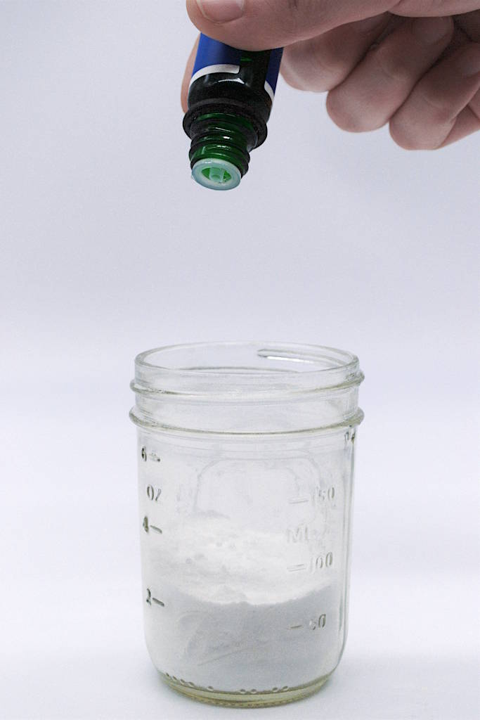 Un-lidded mason jar with baking soda inside with essential oils being added by hand.