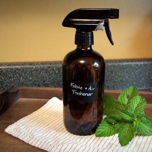 An amber-colored spray bottle labeled Fabric / Air Freshener with some mint leaves beside it sitting on a cloth napkin on a wooden serving tray.