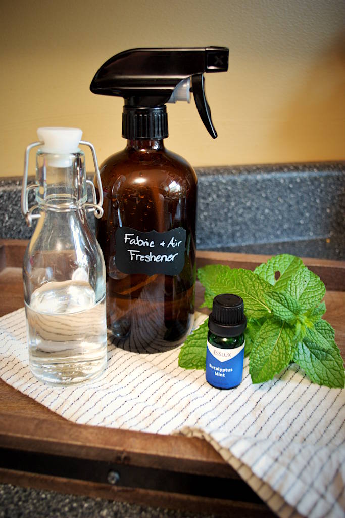 An amber-colored spray bottle labeled Air and Fabric Freshener surrounded by a glass swing-top bottle with vinegar, eucalyptus mint essential oil bottle and mint leaves beside it sitting on a cloth napkin on a wooden serving tray.