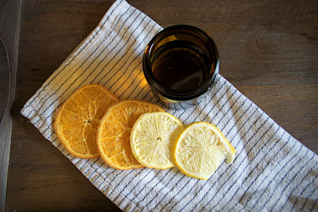 Open amber-colored jar with finished wood polish sitting on a cloth napkin on top of a wooden board. Also on top of the napkin are some freeze-dried orange and lemon slices.