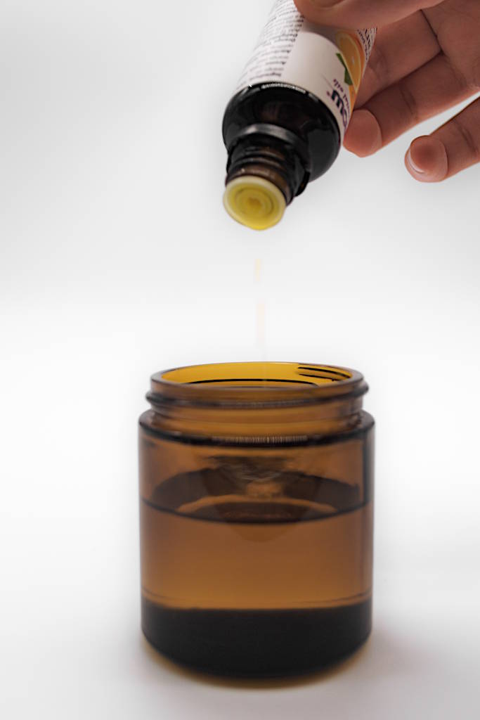 An amber-colored glass jar with oil inside and orange essential oil being added to it.