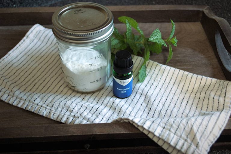 Lidded mason jar with a baking soda and essential oil mixed in sitting on a cloth napkin on a wooden board with a stem of mint leaves behind it and a small bottle of eucalyptus mint essential oil.