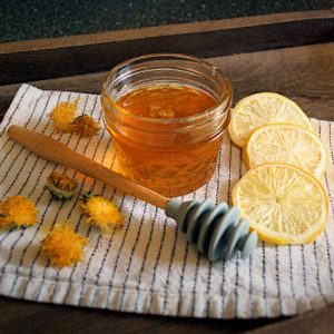 A mason jar with dandelion honey surrounded by freeze-dried lemon slices, dandelion flower heads and a honey dipper.