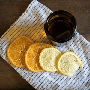 Top view of an open amber-colored glass jar with finished wood polish sitting on top of a cloth napkin surrounded by freeze-dried orange and lemon slices.