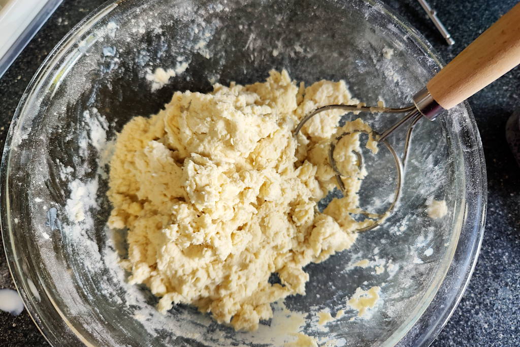 Buttermilk biscuit dough in a glass mixing bowl, mixed up with a Danish dough whisk that is sitting in the bowl.