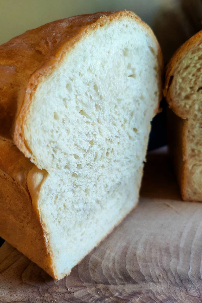 Close up view of a loaf of sandwich bread sitting diagonally on a cutting board that is cut in half to show the crumb.