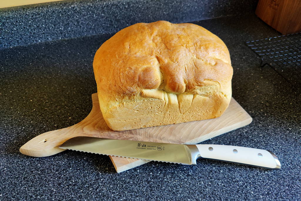 Loaf of sandwich bread sitting on a wooden cutting board with a serrated bread knife.