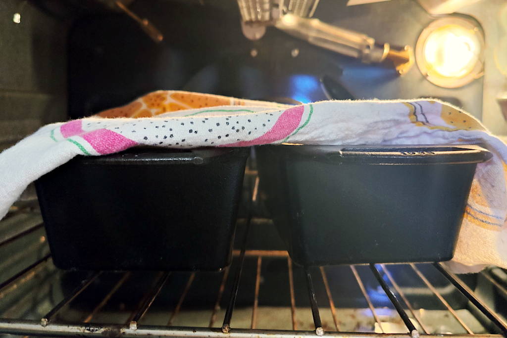 Two cast iron bread pans with a tea towel on top proofing sandwich bread dough in the oven with the light on.