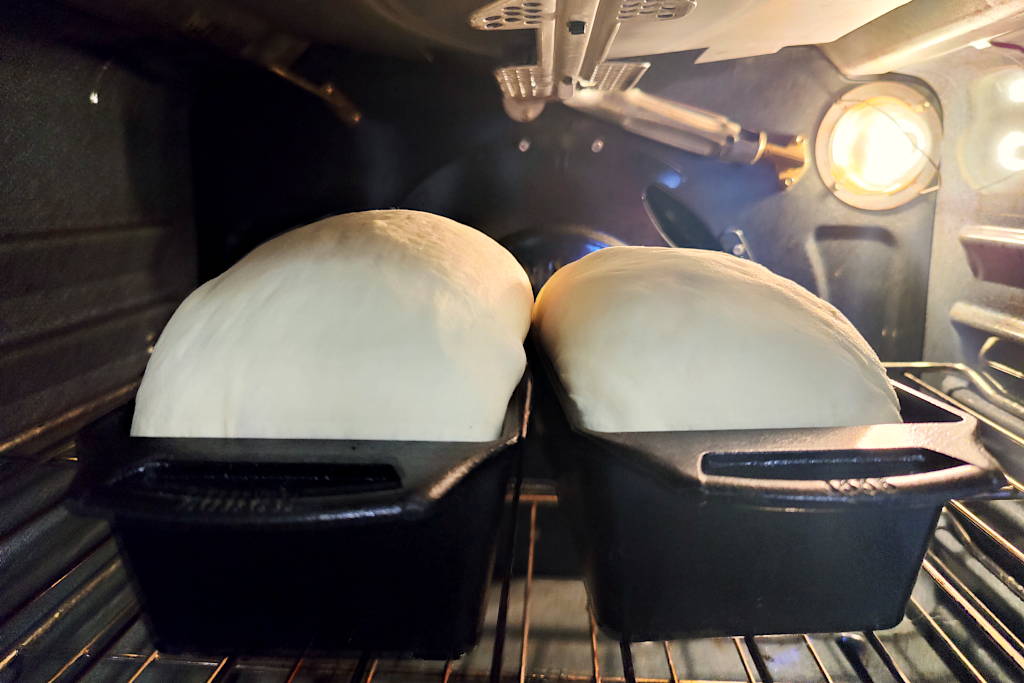 Two loaves of sandwich bread dough inside cast iron bread pans rising in the oven with the light on.